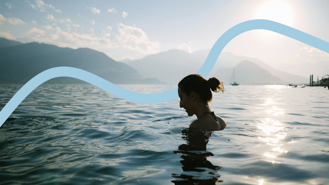 Eine Frau schwimmt bei Sonnenuntergang in einem ruhigen Bergsee. Im Hintergrund sind Berge und Segelboote zu sehen.