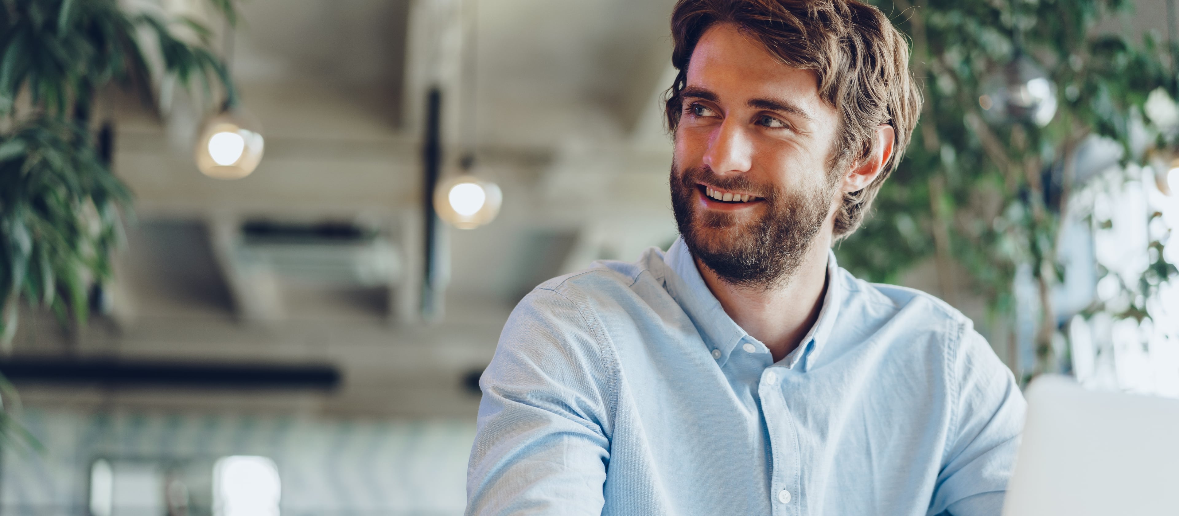 Young man with a beard sits at a desk in a modern, bright office with plants, smiling to the side. A laptop, computer mouse, and documents are placed on the desk in front of him.
