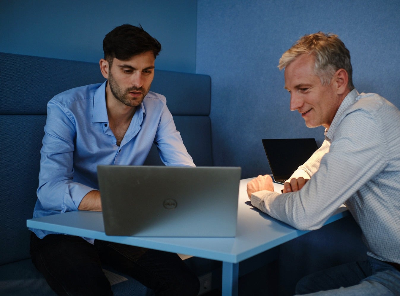 Zwei Kollegen arbeiten gemeinsam an einem Laptop in einer modernen Bürobesprechungsecke.