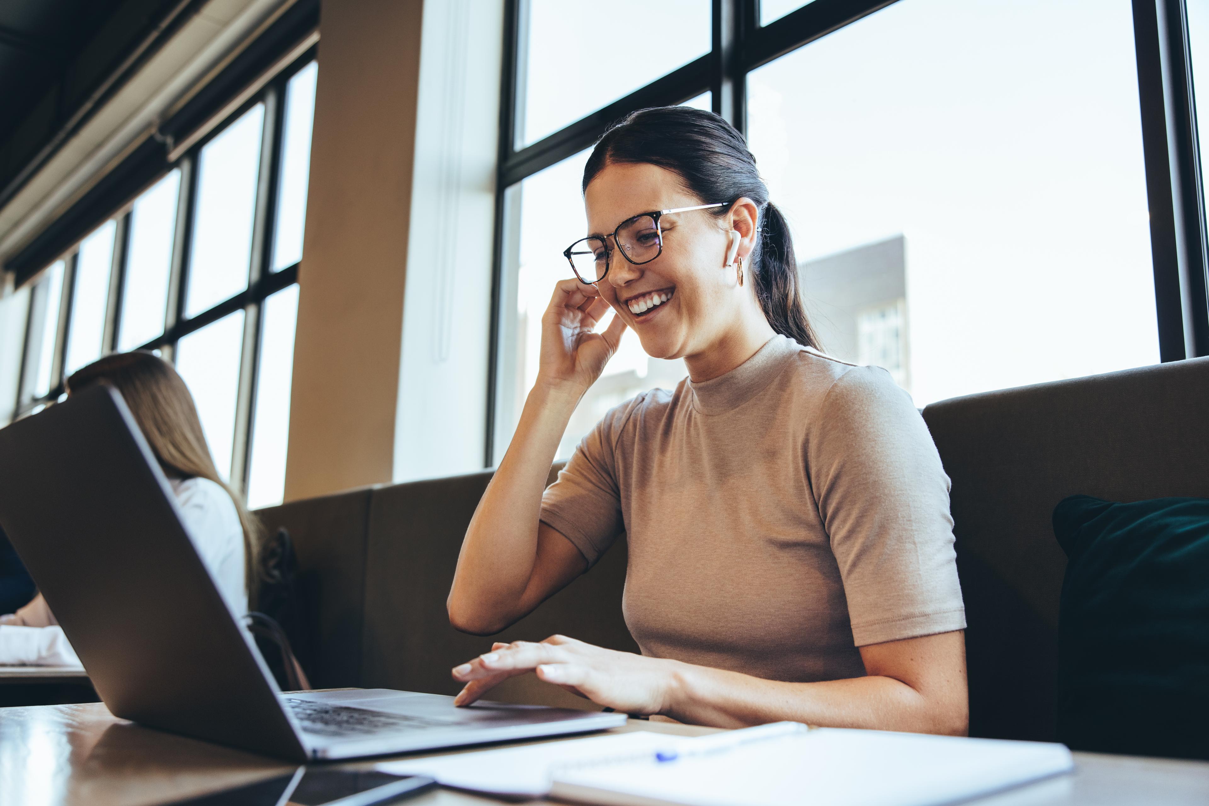 Mavie Work - Mitarbeiterin - Laptop - Open Space Büro Eine junge Frau mit Brille sitzt lächelnd in einem modernen Café oder Co-Working-Space vor ihrem Laptop und telefoniert über Kopfhörer.
