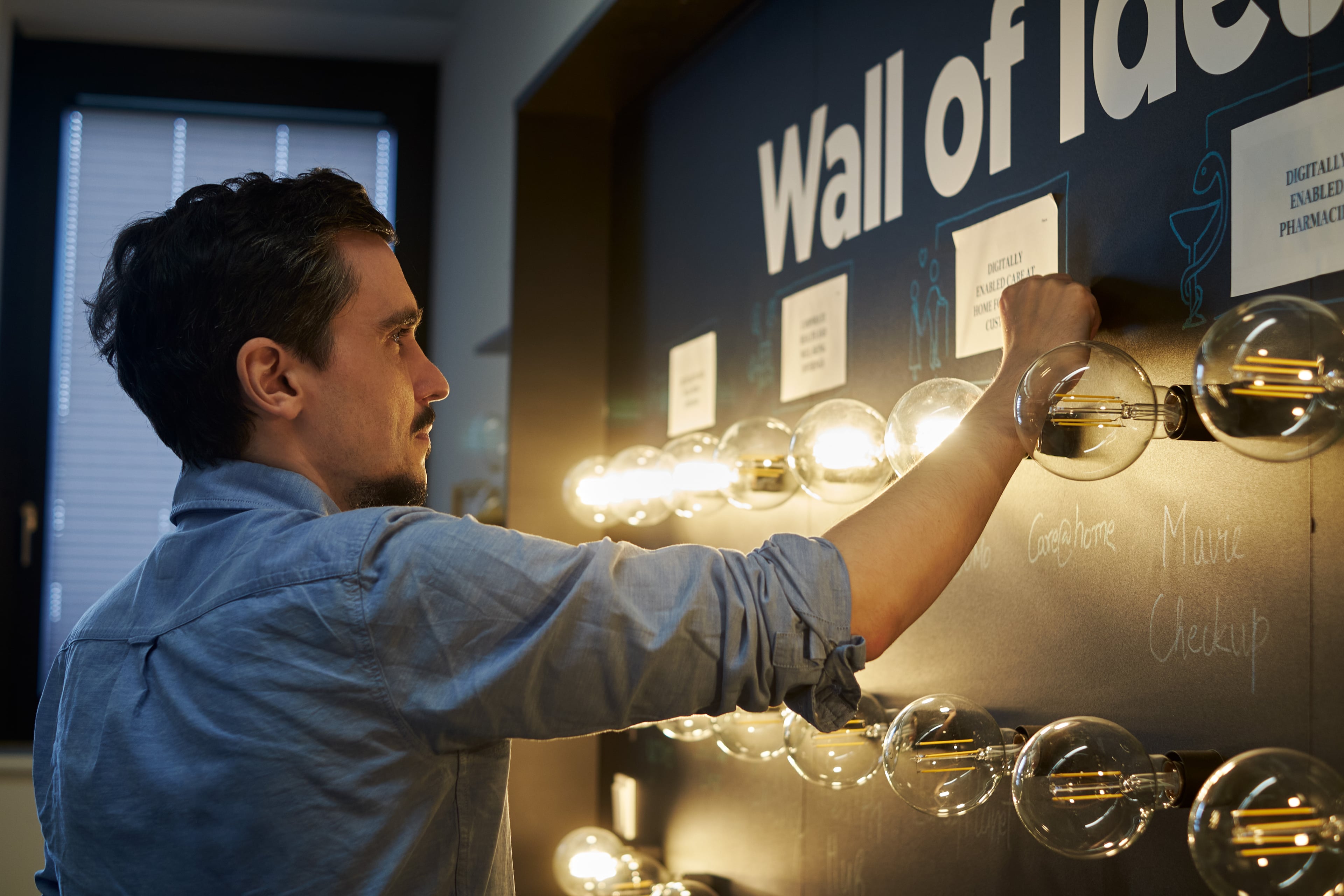 Ein Mann befestigt eine Notiz an einer Wand mit der Aufschrift „Wall of Ideas“, umgeben von dekorativen Glühbirnen – eine kreative Arbeitsumgebung, die Innovation und Ideenaustausch fördert.