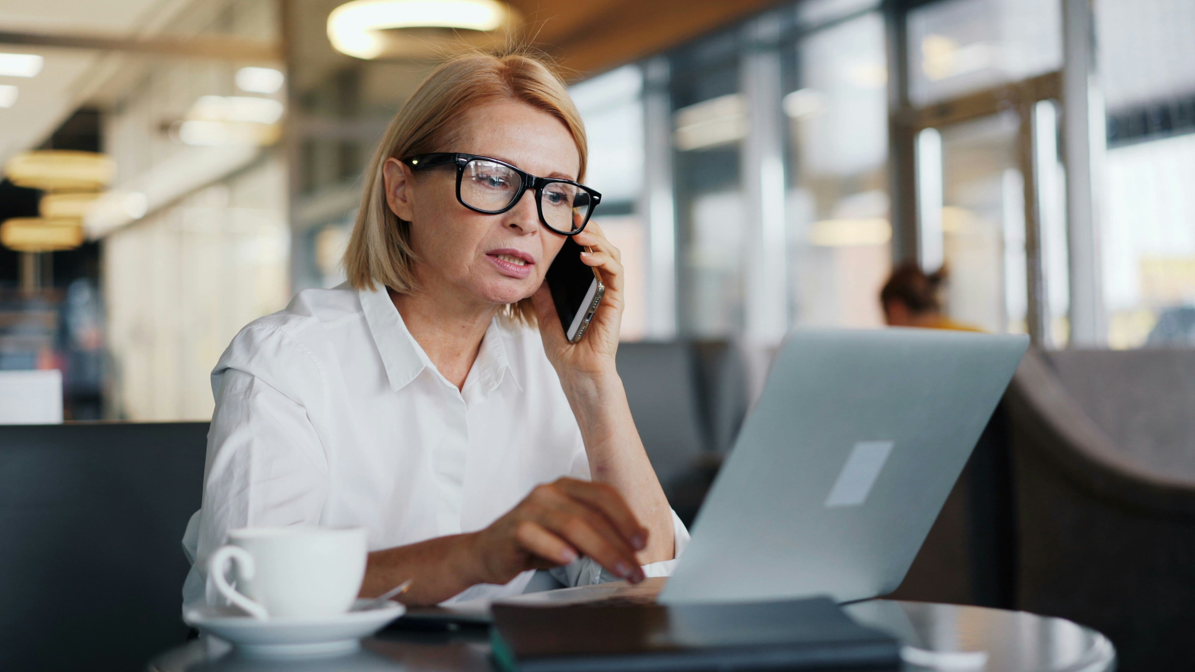 Eine Frau mit blondem Bob und schwarzer Brille sitzt an einem Laptop in einem Café oder Lobby, telefoniert mit dem Smartphone und tippt gleichzeitig, daneben steht eine Kaffeetasse auf dem Tisch.