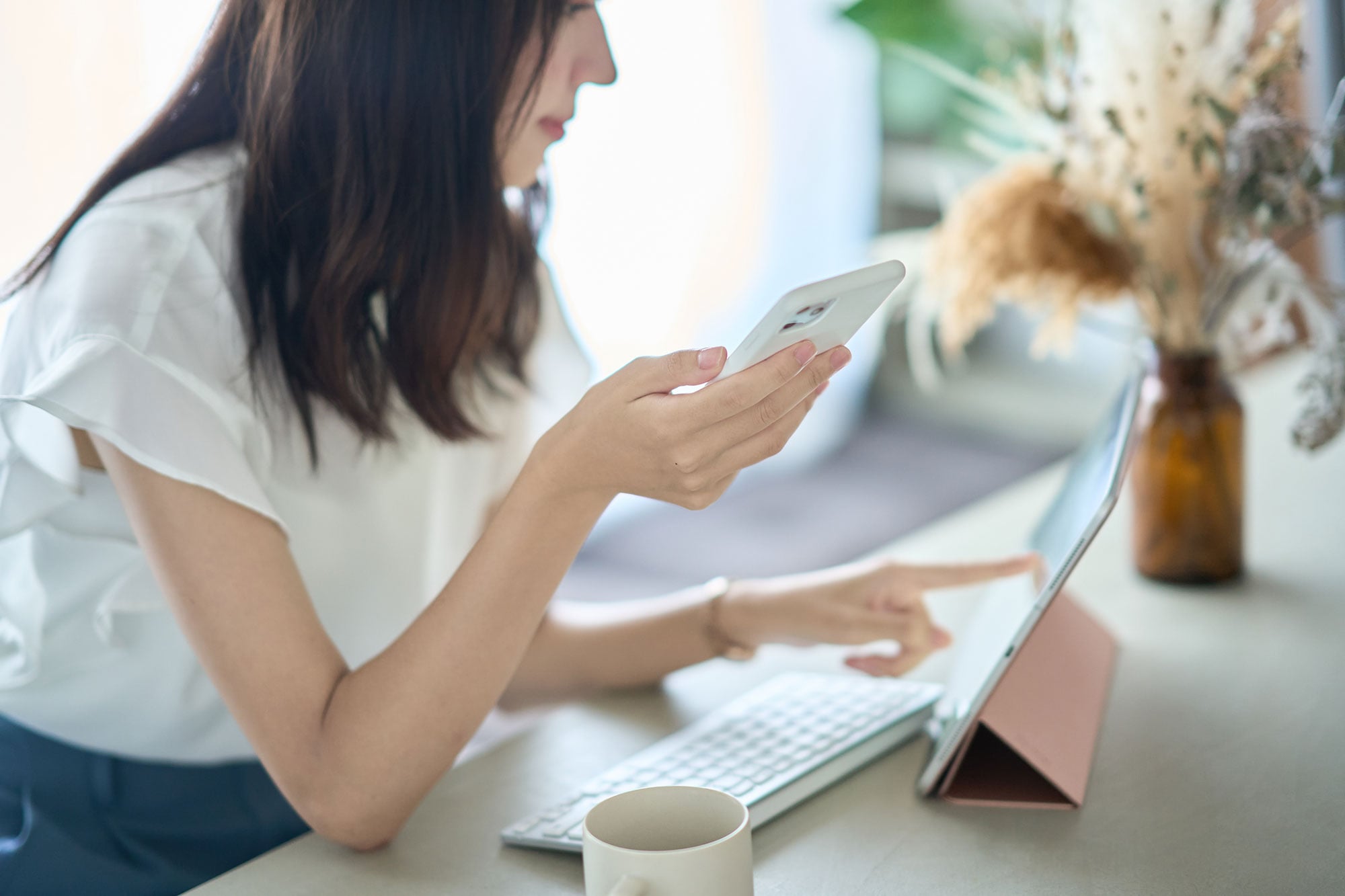 Frau sieht auf ein Tablet und hält gleichzeitig ein Smartphone in der Hand. Auf dem Schreibtisch stehen eine Kaffeetasse und eine Vase mit Trockenblumen.
