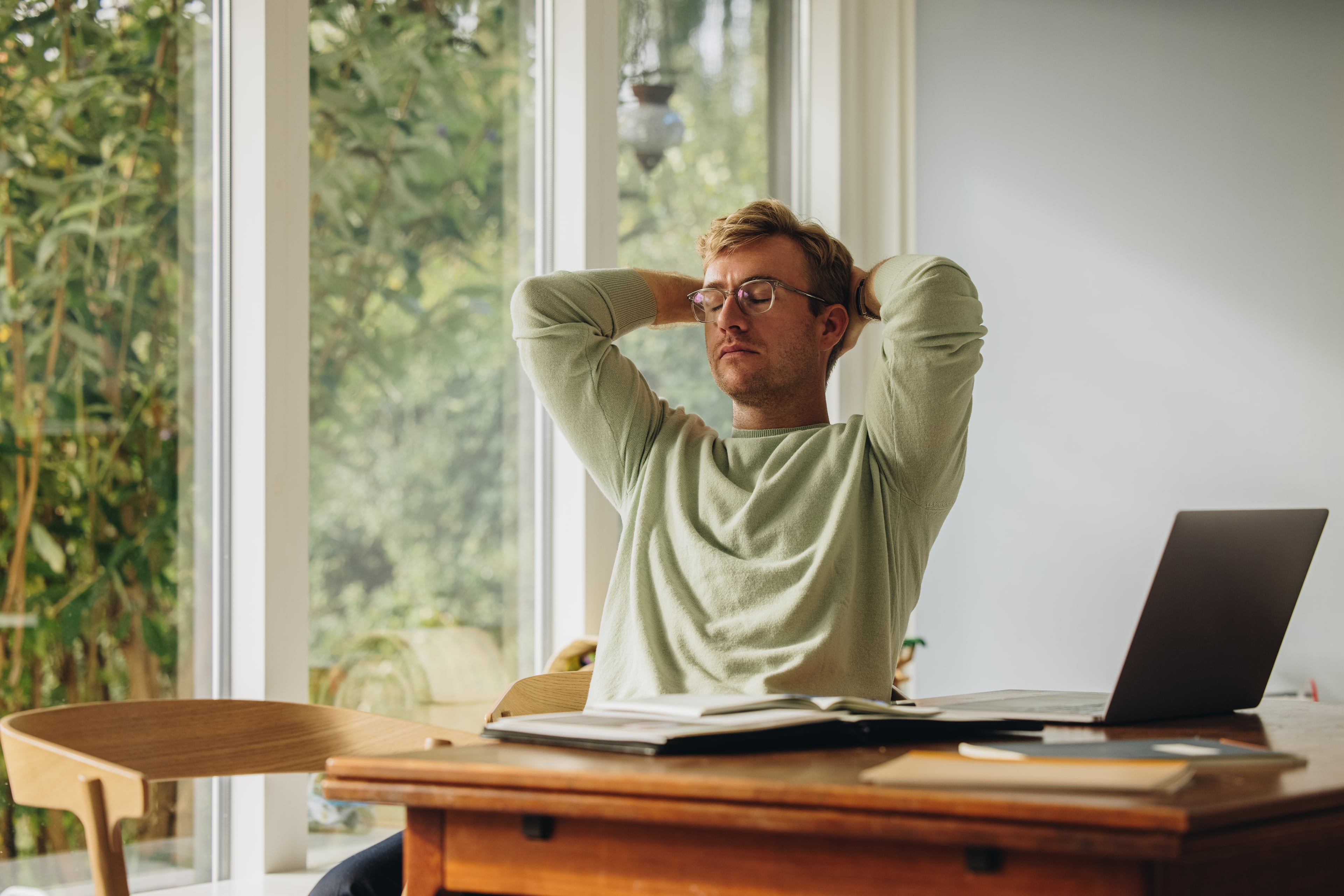 A man wearing glasses sits leaning back with his eyes closed and hands clasped behind his head at a wooden table with open binders and a laptop, large windows with a view of greenery behind him.
