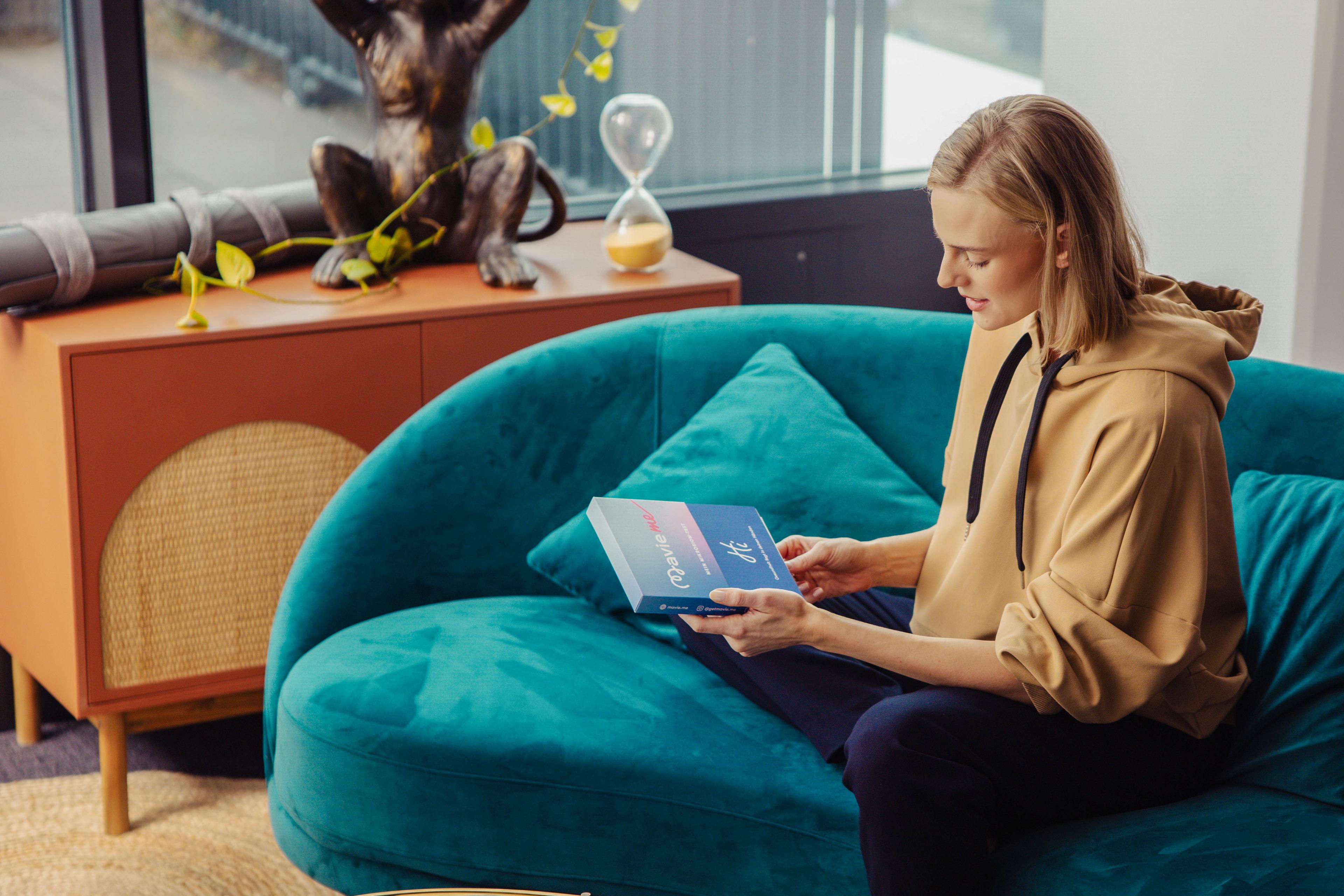 MavieMe Sofa Testkit Eine Frau sitzt entspannt auf einem türkisfarbenen Sofa und hält ein „MavieMe“-Bluttest-Kit in den Händen. Im Hintergrund steht ein Sideboard mit Dekoration.
